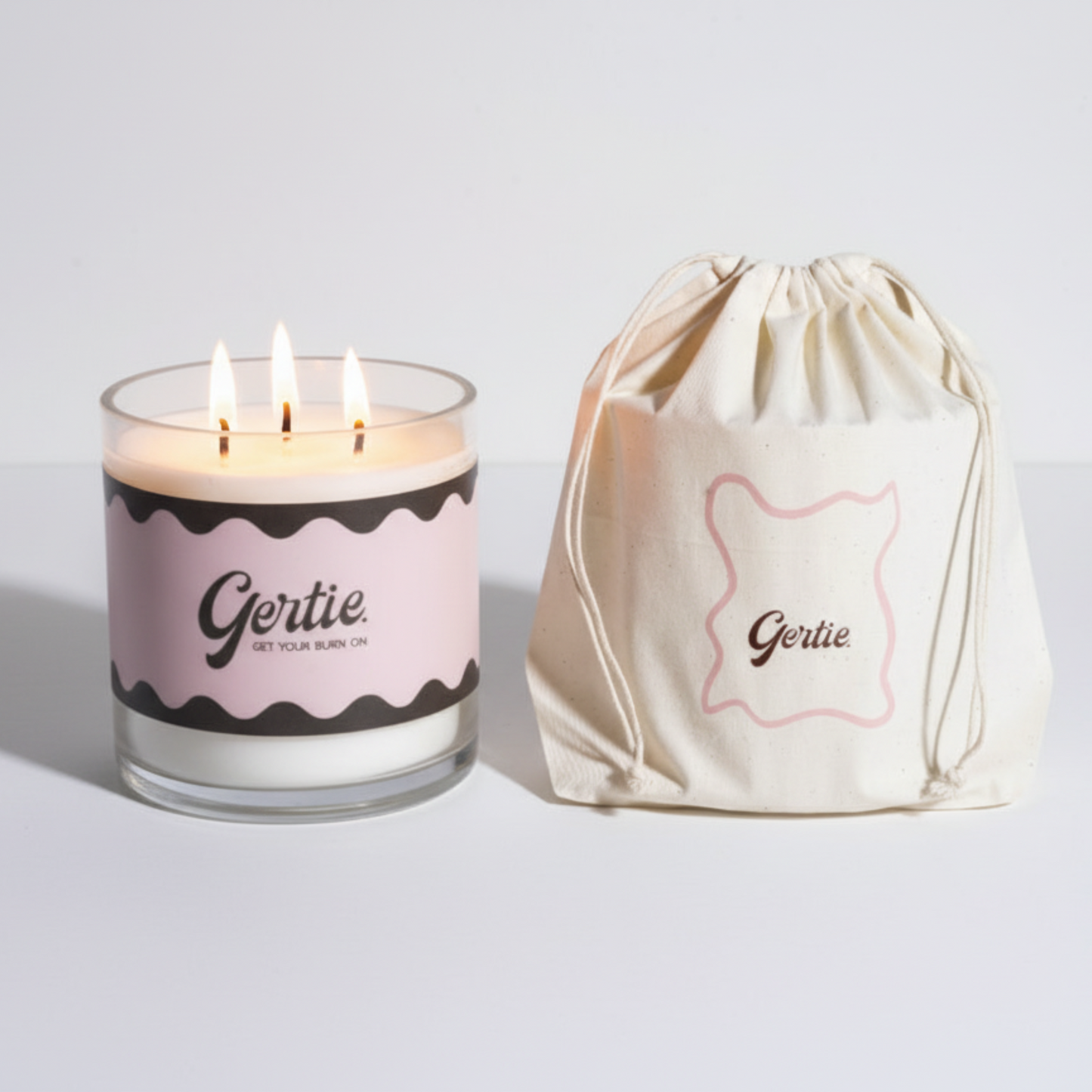 Lit gertie candle with pink and brown label and a matching drawstring bag on a light gray background