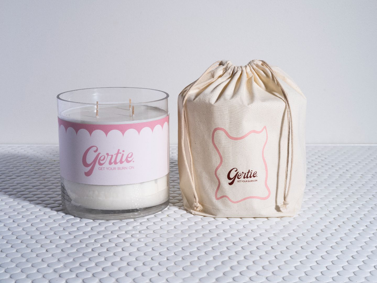A candle with a white wax, pink label, and black writing, alongside a beige drawstring bag with pink writing, both bearing the name 'Gertie'.
