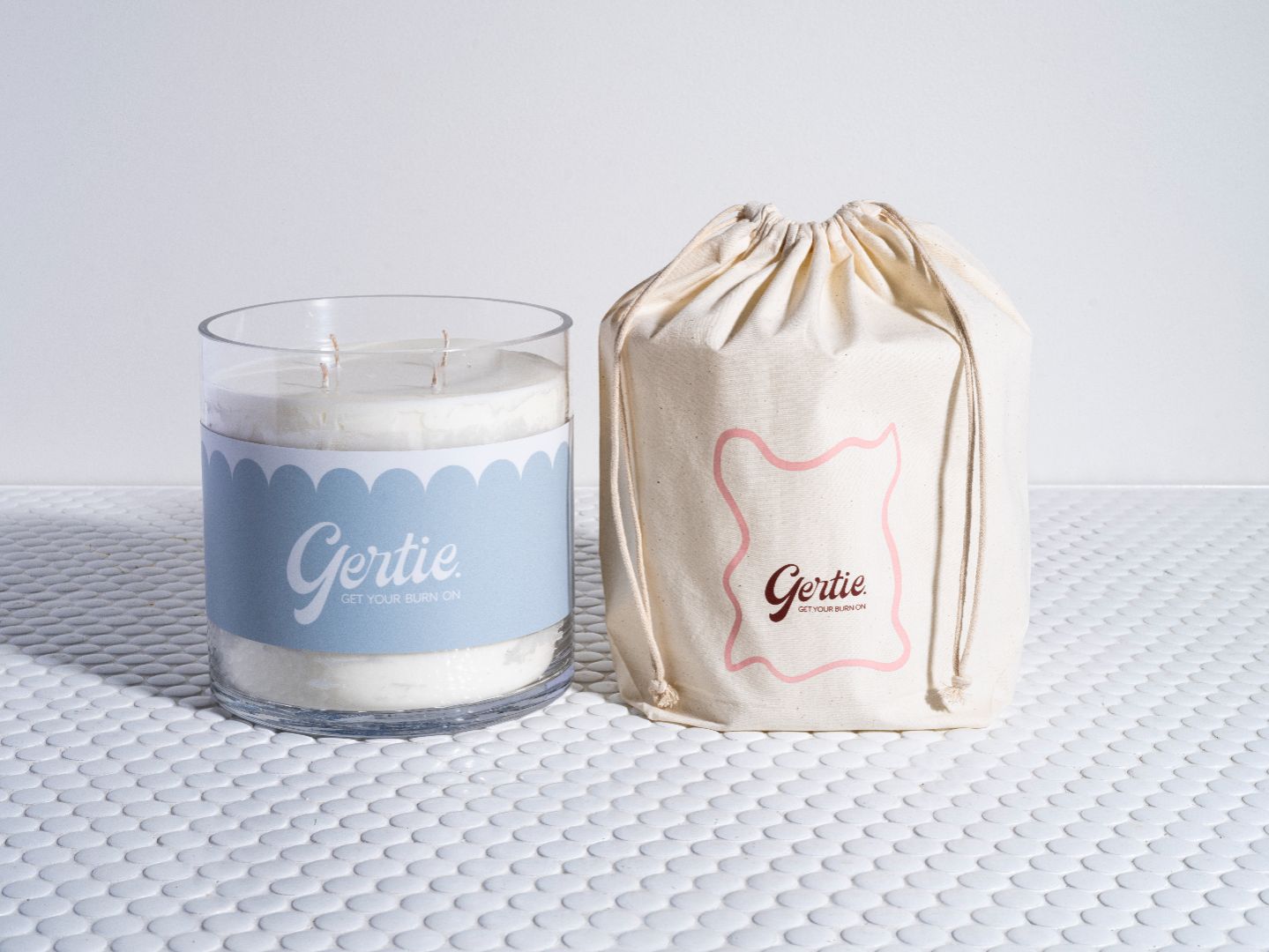 A white candle with a label that reads 'Gertie' and a matching label on a drawstring bag beside it, all presented on a textured surface.