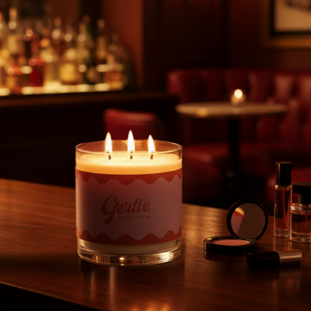 Lit candle with 'Gertie' branding on a bar counter with bottles and makeup.