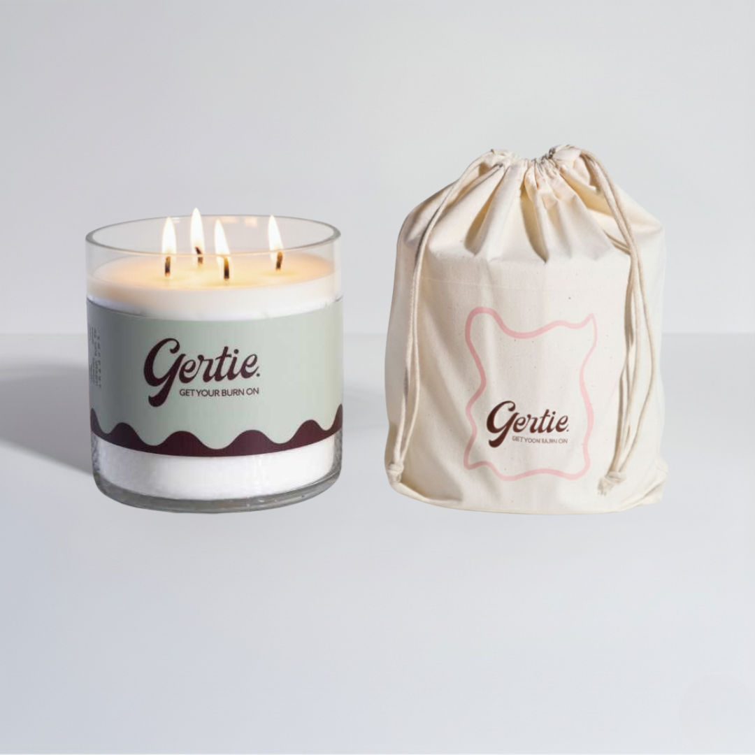Lit gertie candle with green and brown label and a matching drawstring bag on a light gray background