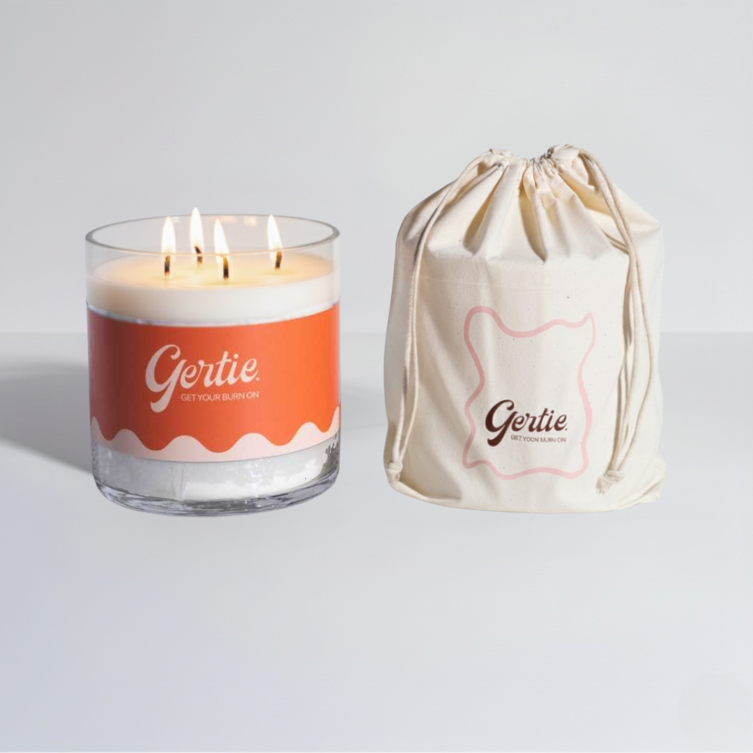 Lit gertie candle with orange label and a matching drawstring bag on a light gray background