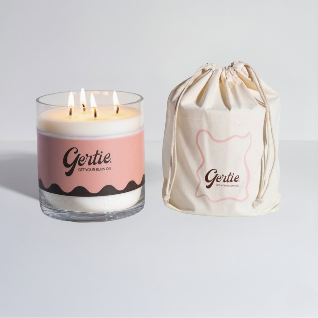 Lit gertie candle with pink and brown label and a matching drawstring bag on a light gray background