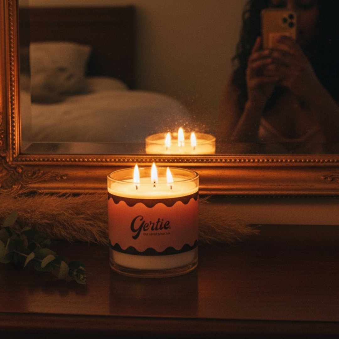 Candle with 'Gertie' label in front of a mirror reflecting a person holding a phone.