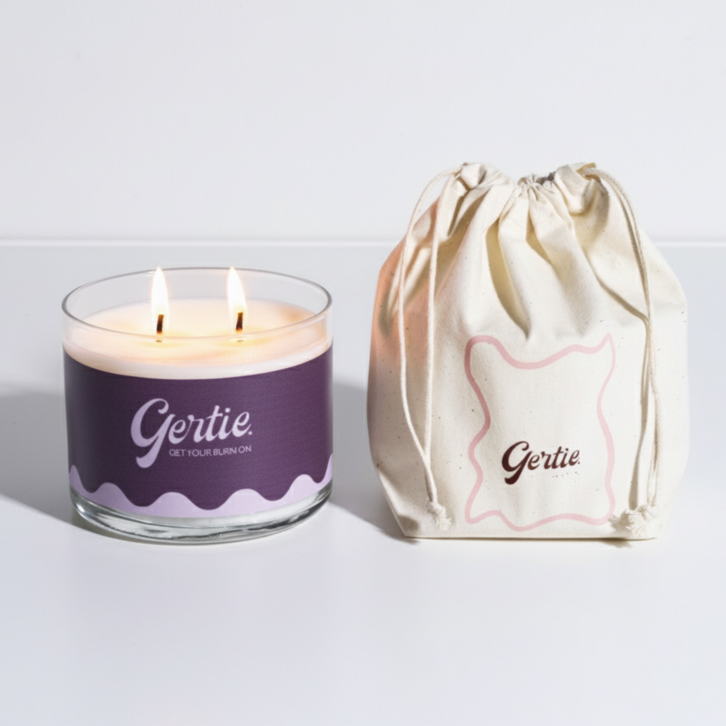 Lit gertie candle with violet label and a matching drawstring bag on a light gray background