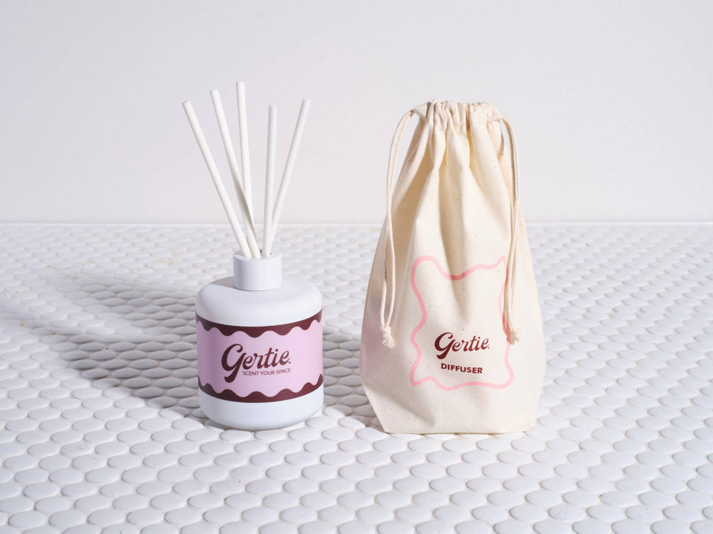 A reed diffuser with white sticks in a white bottle, labeled 'Gertie', next to a beige drawstring bag with the same brand on a textured white surface.