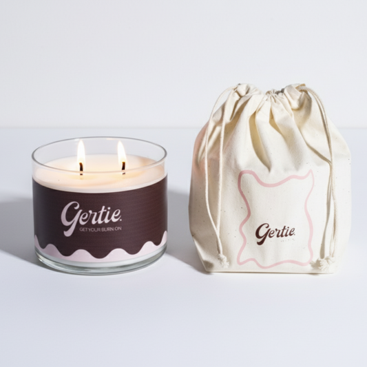Lit gertie candle with brown label and a matching drawstring bag on a light gray background