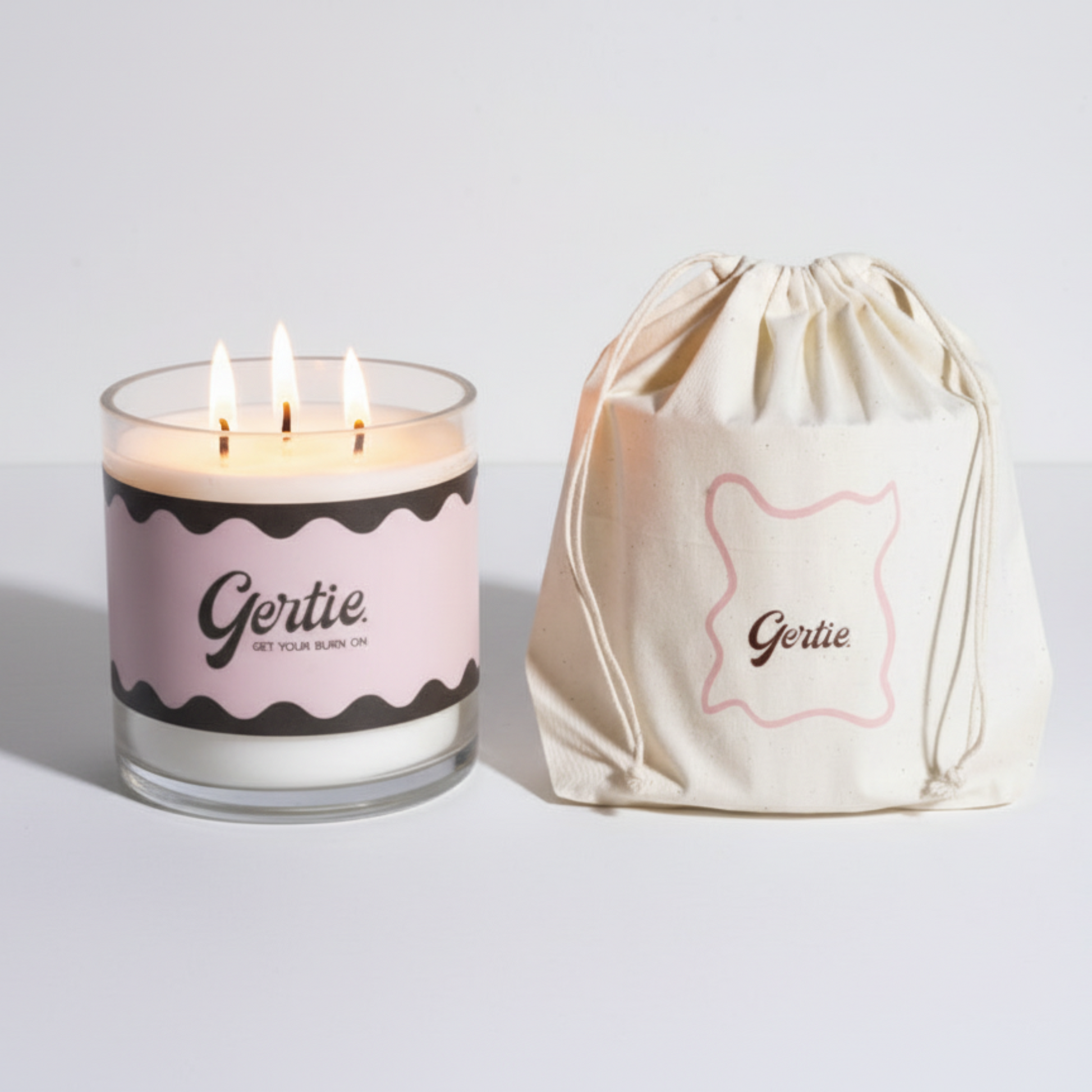 Lit gertie candle with pink and brown label and a matching drawstring bag on a light gray background