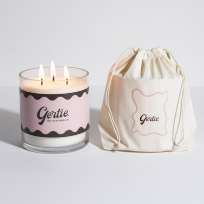 Lit gertie candle with pink and brown label and a matching drawstring bag on a light gray background