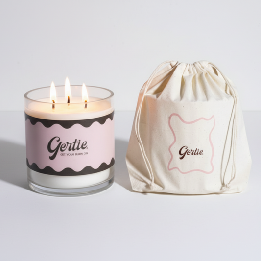 Lit gertie candle with pink and brown label and a matching drawstring bag on a light gray background
