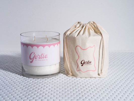 A candle with a white wax, pink label, and black writing, alongside a beige drawstring bag with pink writing, both bearing the name 'Gertie'.