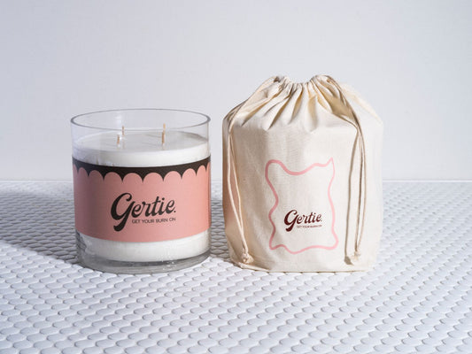 A white candle with a label that reads 'Gertie' and has a pink and black design on it, alongside a beige drawstring bag with the brand name 'Gertie' on it, all presented on a textured white surface.