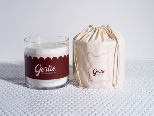 A large candle with a white label and red text, alongside a burlap sack with a red logo, both bearing the brand name 'Gertie'.