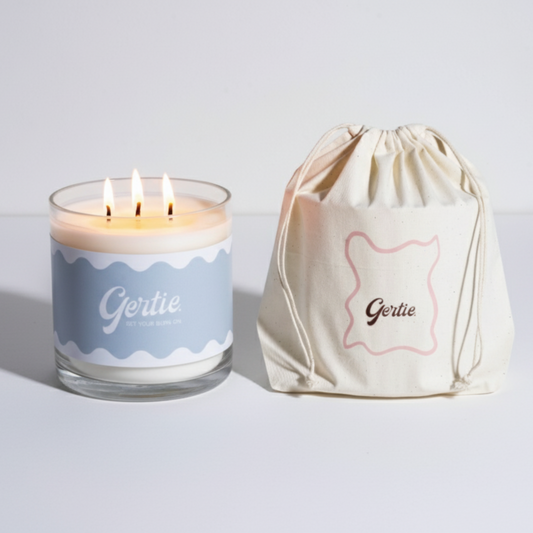 Lit gertie candle with blue label and a matching drawstring bag on a light gray background