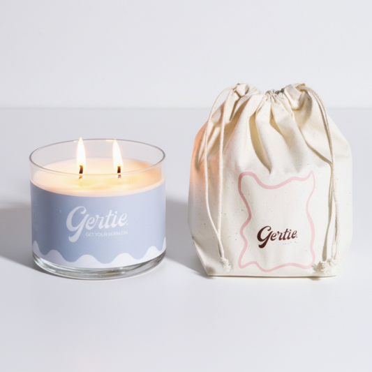 Lit gertie candle with blue label and a matching drawstring bag on a light gray background