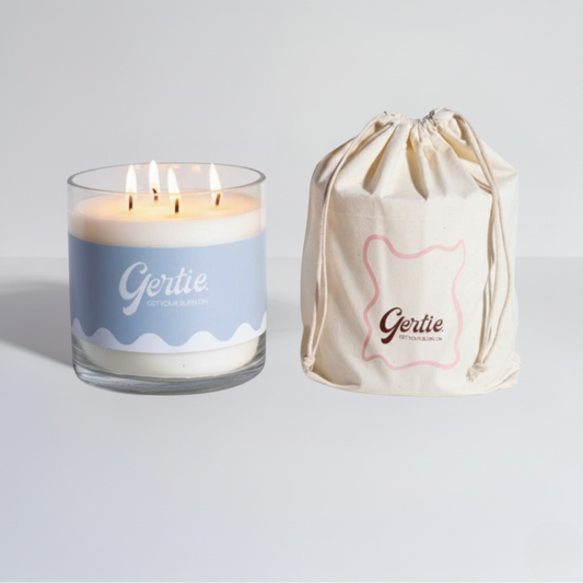 Lit gertie candle with blue label and a matching drawstring bag on a light gray background