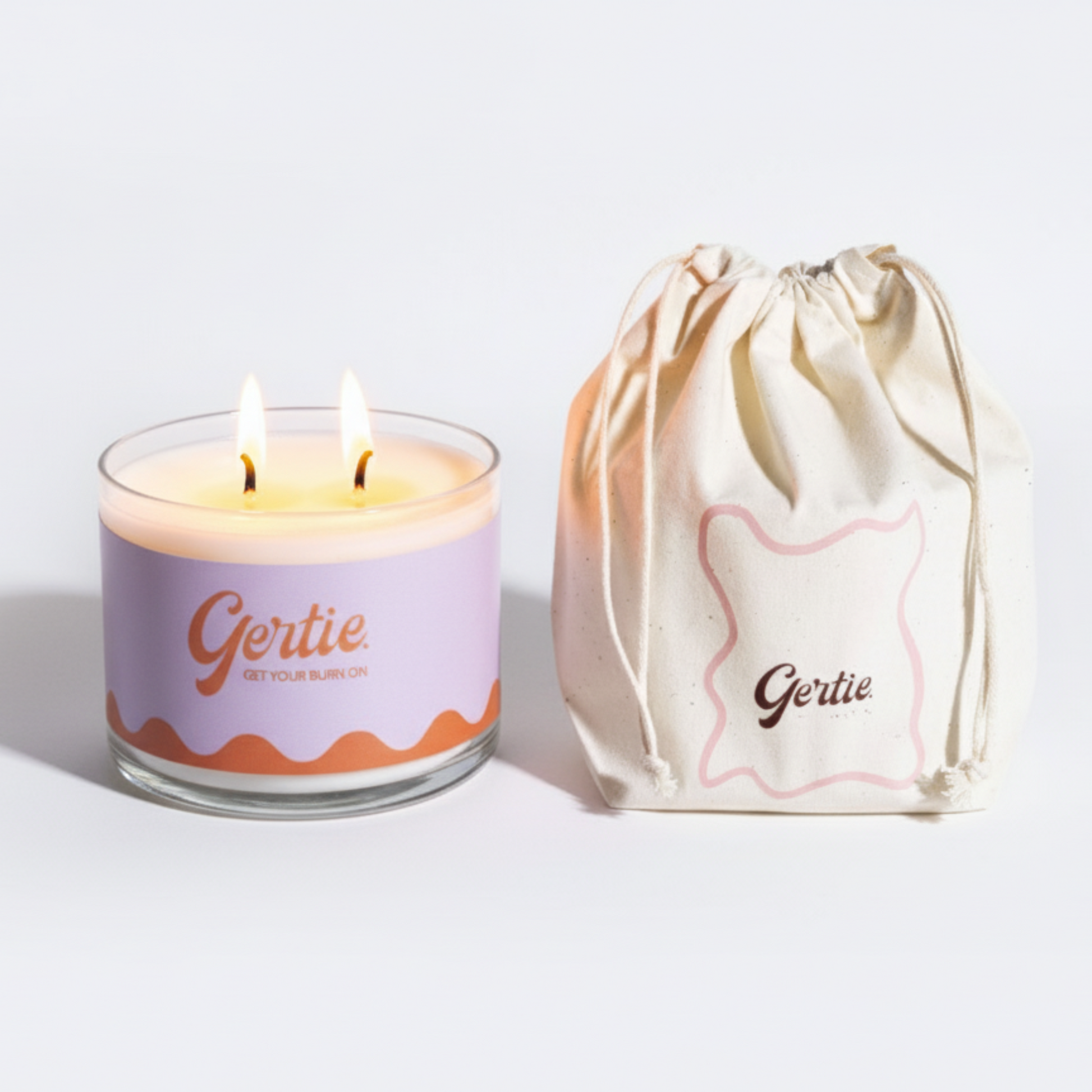 Lit gertie candle with an orange and purple label and a matching drawstring bag on a light gray background