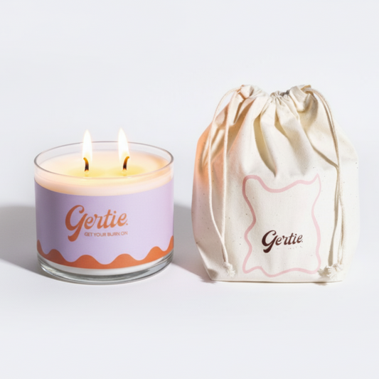 Lit gertie candle with an orange and purple label and a matching drawstring bag on a light gray background