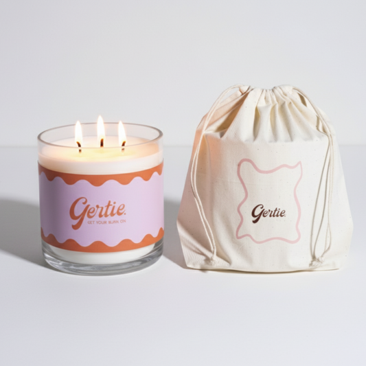 Lit gertie candle with violet label and a matching drawstring bag on a light gray background