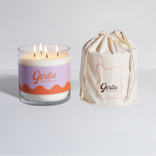 Lit gertie candle with orange and purple label and a matching drawstring bag on a light gray background