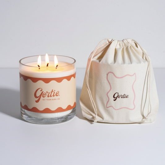 Lit gertie candle with orange label and a matching drawstring bag on a light gray background