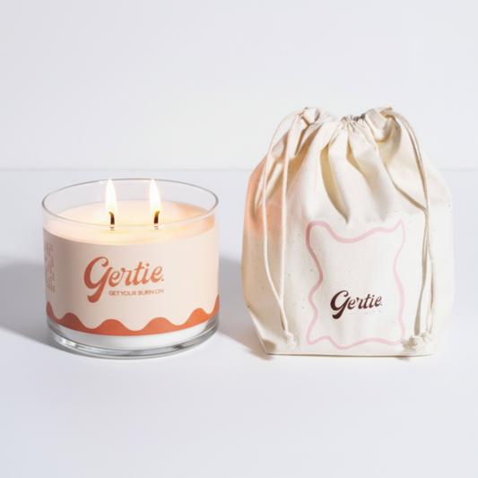 Lit gertie candle with an yellow and orange label and a matching drawstring bag on a light gray background