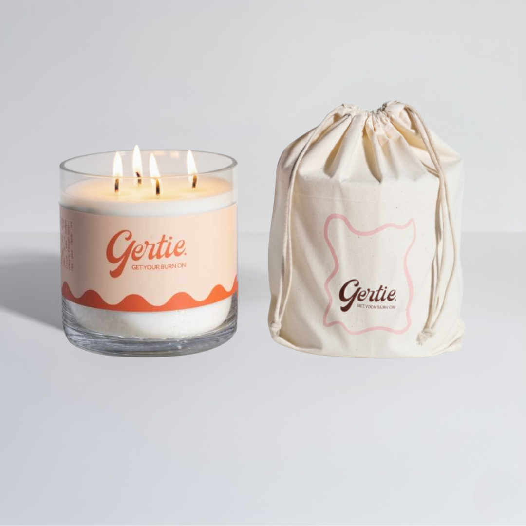 Lit gertie candle with white and orange label and a matching drawstring bag on a light gray background