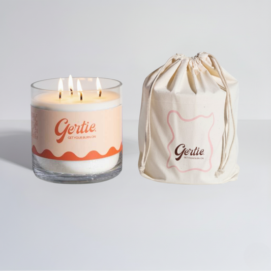 Lit gertie candle with white and orange label and a matching drawstring bag on a light gray background