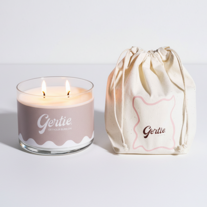 Lit gertie candle with beige label and a matching drawstring bag on a light gray background