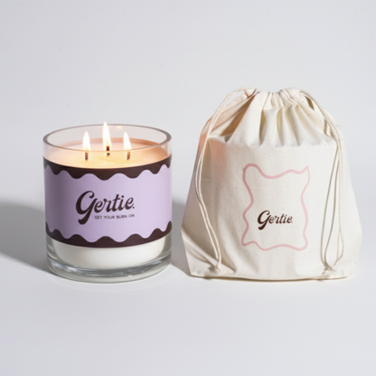 Lit gertie candle with violet label and a matching drawstring bag on a light gray background