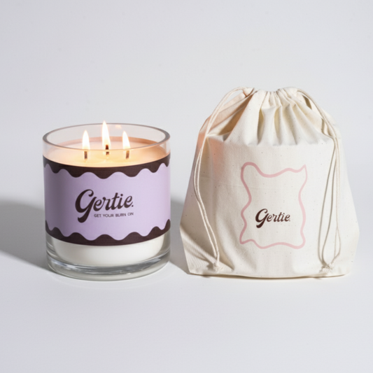Lit gertie candle with violet label and a matching drawstring bag on a light gray background