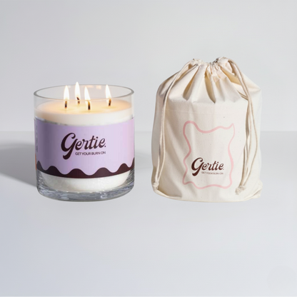 Lit gertie candle with violet and brown label and a matching drawstring bag on a light gray background