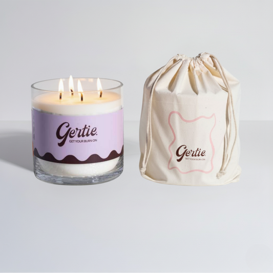 Lit gertie candle with violet and brown label and a matching drawstring bag on a light gray background
