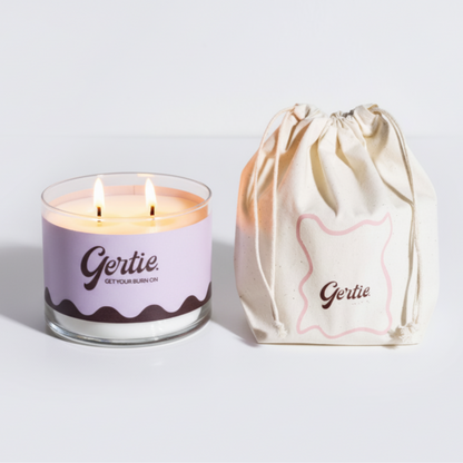 Lit gertie candle with violet label and a matching drawstring bag on a light gray background