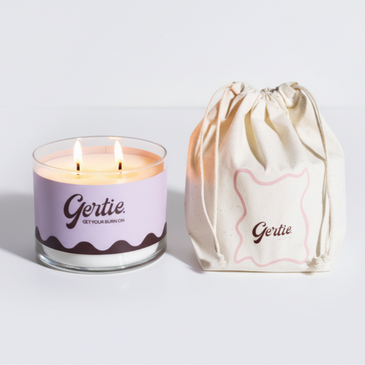 Lit gertie candle with violet label and a matching drawstring bag on a light gray background