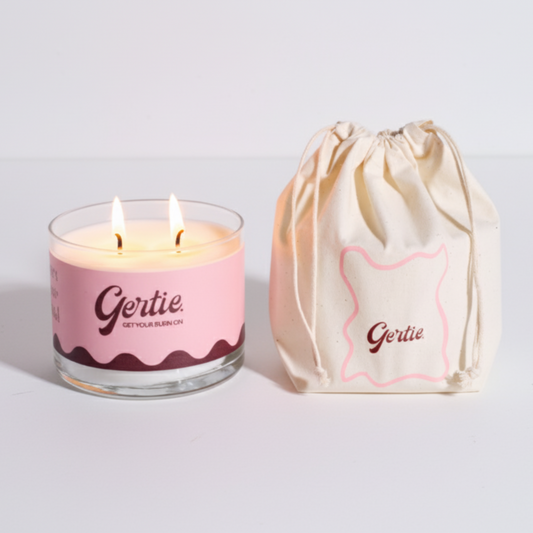 Lit gertie candle with pink label and a matching drawstring bag on a light gray background