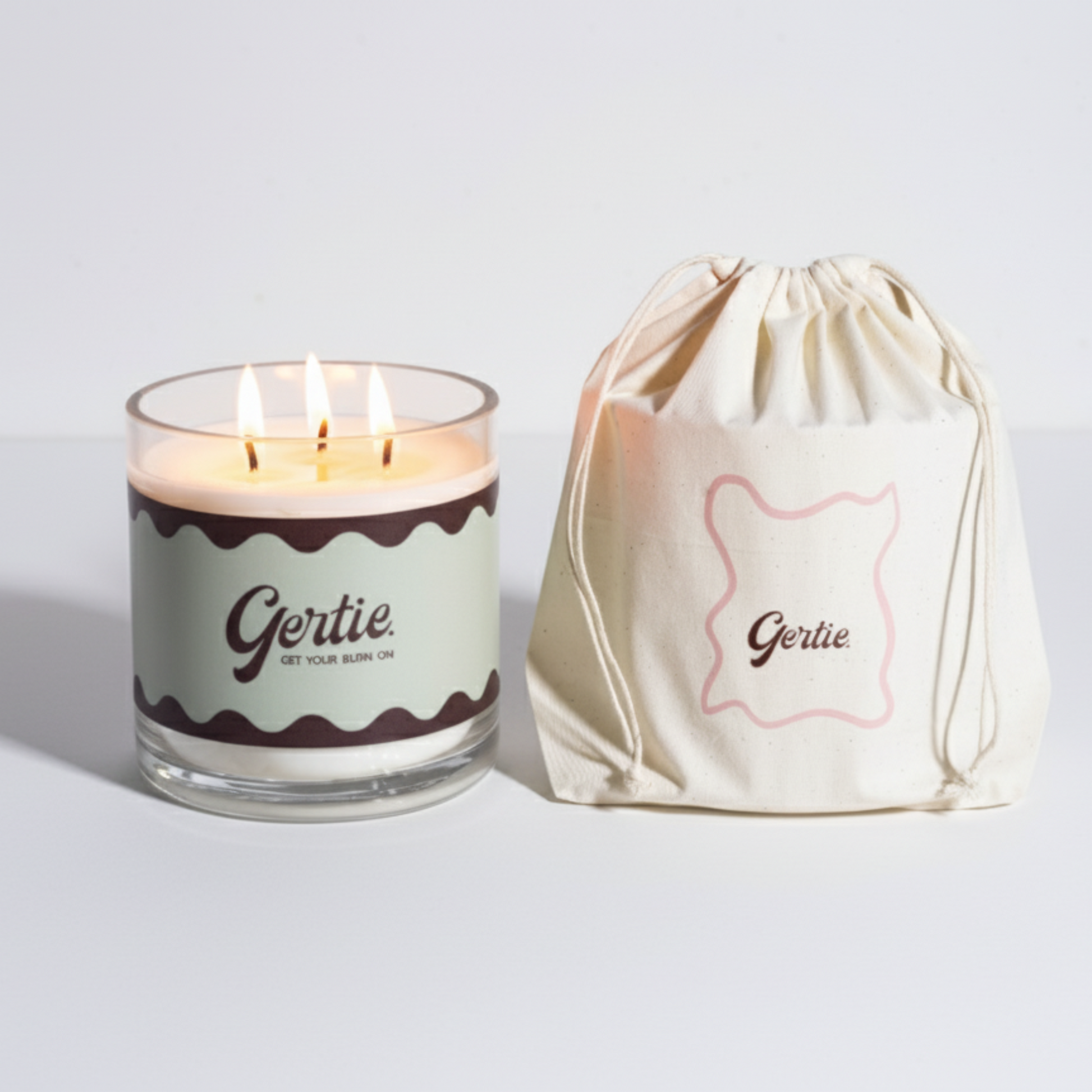 Lit gertie candle with green label and a matching drawstring bag on a light gray background