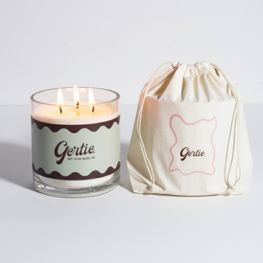 Lit gertie candle with green label and a matching drawstring bag on a light gray background