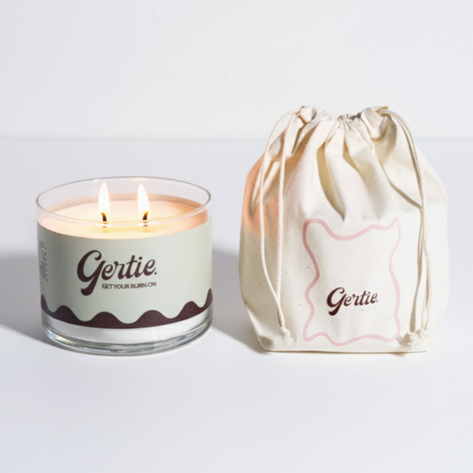 Lit gertie candle with green and brown label and a matching drawstring bag on a light gray background
