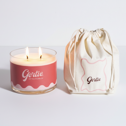 Lit gertie candle with pink label and a matching drawstring bag on a light gray background