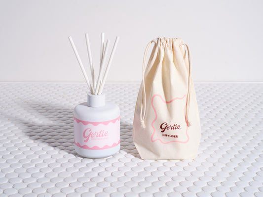 A reed diffuser with white sticks in a white and pink container, accompanied by a beige reusable cotton pouch with the brand name 'Gertie' on it, displayed on a textured white surface.