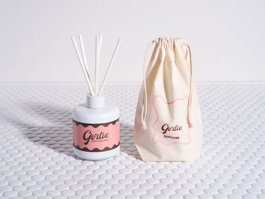 A reed diffuser with white sticks in a white bottle, labeled with 'Gertie' and a peach color, next to a beige cotton pouch with the same brand.