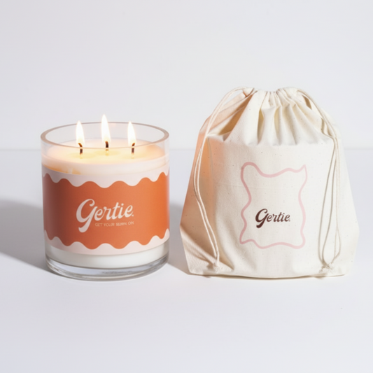 Lit gertie candle with orange label and a matching drawstring bag on a light gray background