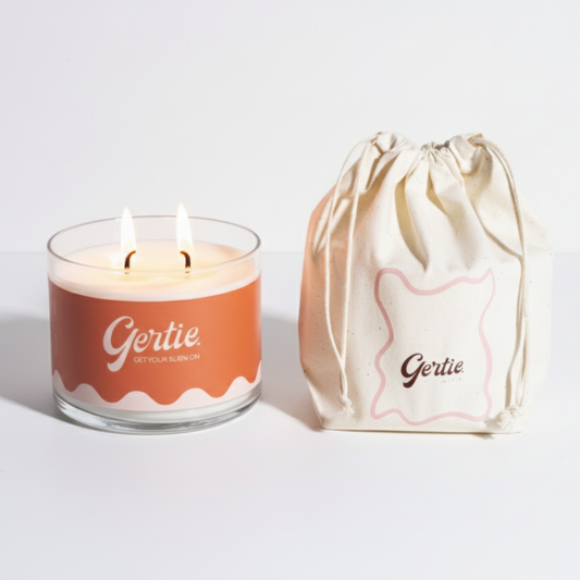 Lit gertie candle with an orange label and a matching drawstring bag on a light gray background