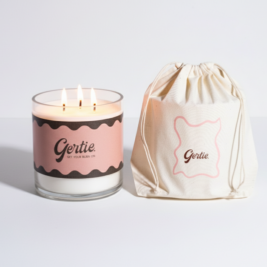 Lit gertie candle with pink label and a matching drawstring bag on a light gray background