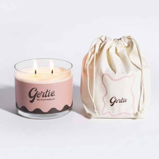 Lit gertie candle with a pink and brown label and a matching drawstring bag on a light gray background