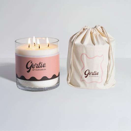 Lit gertie candle with pink and brown label and a matching drawstring bag on a light gray background