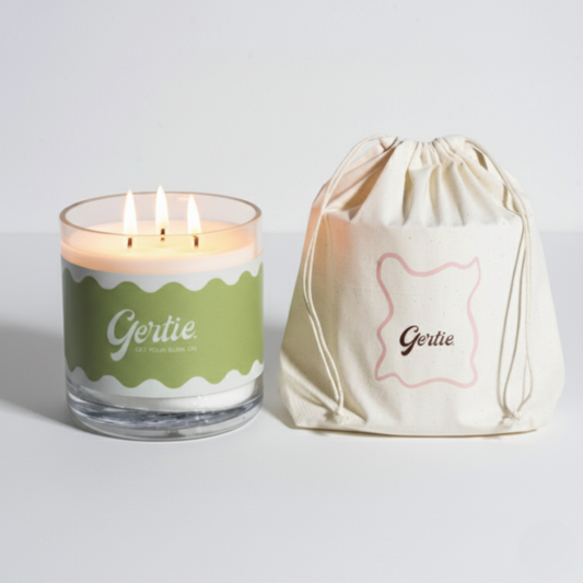 Lit gertie candle with green label and a matching drawstring bag on a light gray background