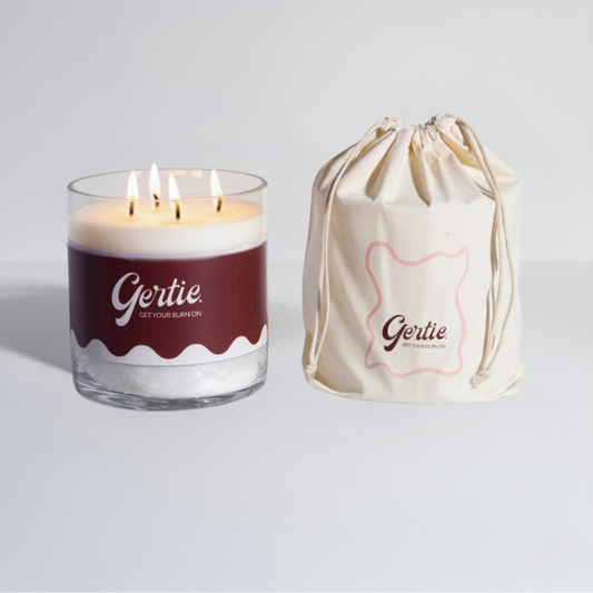 Lit gertie candle with brown label and a matching drawstring bag on a light gray background