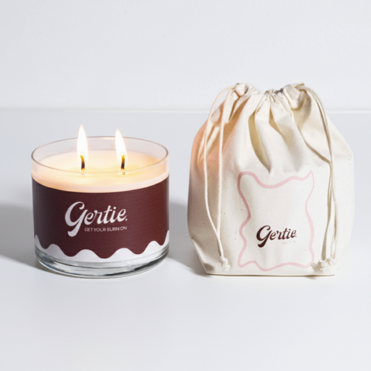 Lit gertie candle with a brown label and a matching drawstring bag on a light gray background